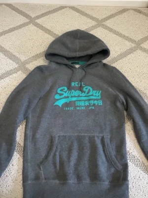 Grå Superdry hoodie med turkost tryck - Snygg grå hoodie från Superdry med turkost logotryck och japansk text på bröstet. Klassisk känguruficka framtill och justerbar huva med dragsko. Perfekt för en avslappnad streetwear-look. Mjuk bomullsblandning för extra komfort.