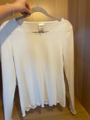 Vit långärmad topp från Kappahl - Söt vit långärmad topp från Kappahl med liten rosett vid halsringningen och vågig kant nertill. Toppen är ribbad och har en klassisk, enkel design som passar till mycket. Perfekt att styla med jeans eller kjol.pris kan diskuteras!💗