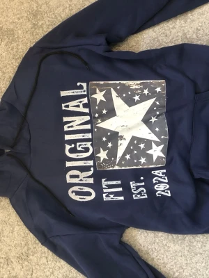 Mörkblå hoodie med stjärntryck - Snygg mörkblå hoodie från Original Fit med stor vit stjärna och flera mindre stjärnor på bröstet. Trycket har en vintage-look och texten 'ORIGINAL FIT EST. 2021' i vitt. Hoodien har huva och känguruficka framtill. Perfekt för en avslappnad streetstyle. Säljer den för att den inte passar. (Rep) Storleken är L men den är egentligen en M. Om du brukar ha M så passar denna perfekt skulle jag tro.