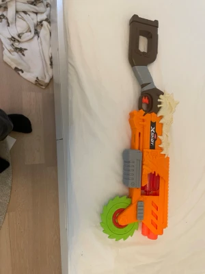 X-Shot leksaksmotorsåg cosplay - Cool leksaksmotorsåg från X-Shot, perfekt för maskerad eller cosplay. Sågen är orange med grönt sågblad, gråa detaljer och ett handtag i brunt. På ovansidan sitter en dekorativ benliknande detalj som ger ett extra tufft intryck.