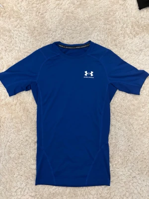 Blå kompressionströja Under Armour - Säljer en blå kompressionströja från Under Armour med korta ärmar och rund hals. Tillverkad i ett stretchigt funktionsmaterial (HeatGear) som andas och passar perfekt till träning. Logga i vitt på bröstet.