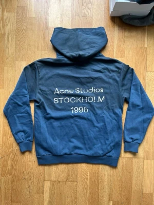 Blå hoodie från Acne Studios - Säljer en blå hoodie från Acne Studios med trycket 'Acne Studios STOCKHOLM 1996' i vitt på ryggen. Hoodien har klassisk huva, ribbade muddar och är gjord i mjukt bomullsmaterial. Perfekt för dig som gillar streetwear och vill ha något stilrent men ändå unikt.