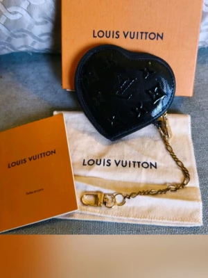 Svart hjärtformad coin purse  - Snygg hjärtformad coin purse i svart lackat läder med klassiskt monogram. Den har en guldfärgad kedja och karbinhake, perfekt att fästa i väskan eller nyckelringen. Kommer med originalförpackning och dustbag.