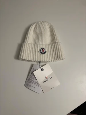 Vit ribbstickad mössa från Moncler - Säljer en stilren vit ribbstickad mössa från Moncler med uppvikt kant och Monclers klassiska logotyp framtill. Mössan är tillverkad i mjukt material och har en clean look som passar perfekt till kalla dagar.
