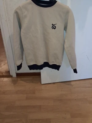 Beige sweatshirt med mörkblå detaljer - Säljer en beige sweatshirt med mörkblå ribbade muddar vid hals, ärmslut och nederkant. Tröjan har en liten logga på bröstet och är tillverkad i mjukt material. Perfekt för en avslappnad stil.