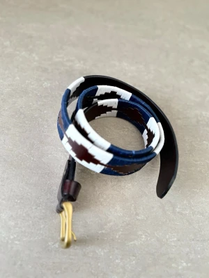 Brunt och blått mönstrat läderbälte - Snyggt bälte i brunt skinn med coolt geometriskt mönster i blått och vitt. Bältet har en klassisk guldfärgad metallspänne och passar perfekt för att lyfta vilken outfit som helst.