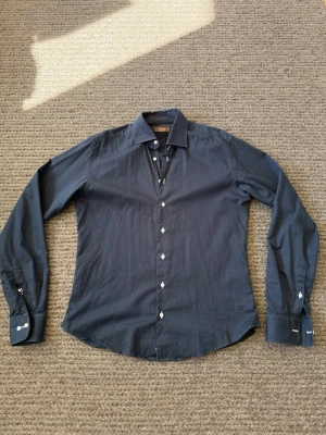 Mörkblå skjorta från iTailor - Stilren mörkblå skjorta från iTailor med klassisk krage och vita knappar. Skjortan har lång ärm och är tillverkad i ett mjukt bomullstyg som känns skönt mot huden. Perfekt för dig som gillar en clean och enkel look.