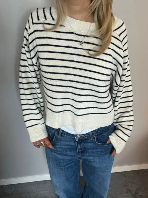 randig stickad tröja - Snygg stickad tröja i vitt med svarta horisontella ränder. Tröjan har en avslappnad passform, rund halsringning och ribbade muddar vid ärmslut och nederkant. Perfekt att matcha med jeans för en stilren look.