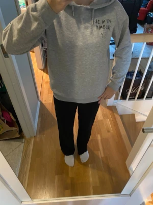 Grå hoodie med text 'JE M'EN AMOUR' - Säljer en grå hoodie med huva och dragsnören. På bröstet och ryggen står det 'JE M'EN AMOUR' i svart text och en stjärna är ritad under texten på ryggen. Hoodien har en klassisk passform och ribbade muddar vid ärmar och nederkant. Kan vara lite trasig vid halsen där luvan sätts ihop. Kontakta mig vid fråga.