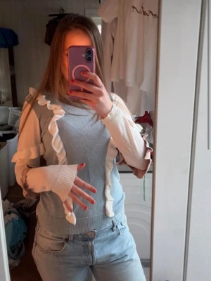Grå stickad tröja med volangdetaljer - Supersöt grå stickad tröja med vita volangdetaljer längs ärmar och framstycke. Tröjan har långa ärmar och breda muddar med tumhål. Perfekt för dig som gillar unika detaljer och en lekfull stil.
