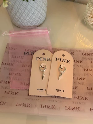 Silvriga örhängen från PINK BOW19 - Säljer ett par silvriga örhängen från PINK BOW19 med en cool, spetsig hängande detalj. Örhängena har en minimalistisk design och kommer med originalförpackning och rosa påse. 