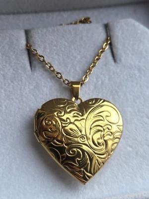 Guldfärgat medaljong hjärthalsband med mönster - Snyggt halsband i guldfärgat rostfritt stål med ett hjärtformat medaljonghänge. Medaljongen har ett detaljerat, blommigt mönster på framsidan och kan öppnas för att lägga in ett foto eller minne. Kedjan är klassisk och matchar hängets eleganta look. Perfekt accessoar för dig som gillar romantisk stil.