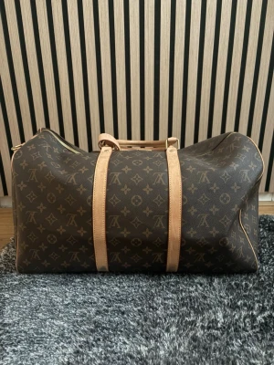 Louis Vuitton Keepall Monogram väska (ej äkta) - Säljer en klassisk Louis Vuitton Keepall resväska i brunt monogramcanvas med ljusbruna läderdetaljer och guldfärgade metalldragkedjor. Väskan har dubbla handtag och ikoniskt LV-mönster över hela ytan. Perfekt för weekendresor och riktigt snygg att bära.