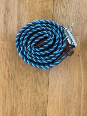 Randigt blått och grått bälte - Snyggt randigt bälte i blått och grått med detaljer i brunt skinn och ett silvrigt spänne. Bältet har en flätad design som ger en cool och modern look. Perfekt för att lyfta vilken outfit som helst.