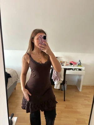 Brun volangklänning med spetsdetaljer - Söt brun klänning med smala axelband och spetsdetaljer vid urringningen. Klänningen har volangkant nertill och sitter figurnära upptill. Perfekt för dig som gillar en feminin och trendig stil. Vet ej om detta klassas som ett linne,nattlinne eller klänning men man kan använda den utifrån sin egen fantasi 🥰defekt:lite nopprig!