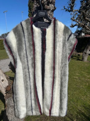 Randig pälsväst från Rodebjer - Snygg pälsväst från Rodebjer fw 2014 i grått, vitt och vinrött. Västen har raka, vertikala ränder och är ärmlös med öppen front. Tillverkad i mjuk fuskpäls som ger en lyxig känsla och passar perfekt för att lyfta din outfit.