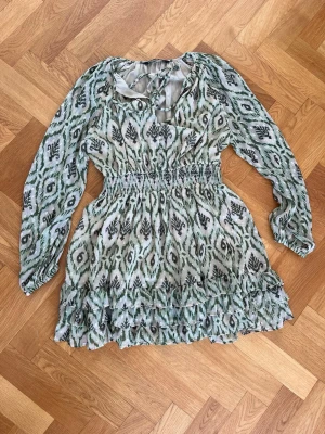 Grönmönstrad kortklänning med volang - Säljer en kort klänning med grönt och svart mönster, v-ringning med snörning och långa ärmar. Klänningen har smock i midjan och volangdetaljer nertill. Tunt, luftigt material som passar perfekt till varmare dagar.