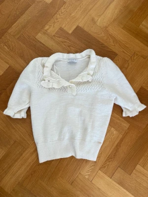 Vit stickad topp med volangkrage - Söt vit stickad topp från Lindex med korta puffärmar och volangdetaljer vid krage och ärmslut. Tröjan har dekorativa knappar framtill och ett diskret mönster upptill. 