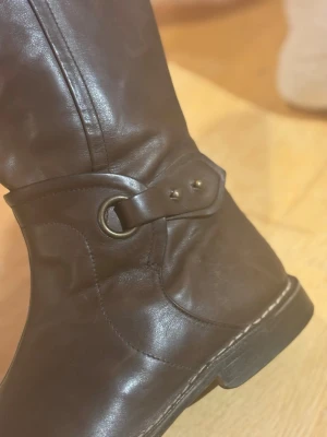 Bruna skinnstövlar från Gidigio - Snygga bruna stövlar från Gidigio, tillverkade i Italien av äkta skinn. Modellen har rund tå, platt sula och dekorativ rem med metallring och nitar vid ankeln. Klassisk design med diskreta sömmar och högt skaft som ger en stilren look.