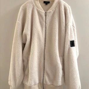 Beige teddyjacka från smog  - Mysig beige teddyjacka från smog med dragkedja framtill och ribbade muddar. Jackan har en avslappnad passform och en svart patch med logga på ärmen. Perfekt för dig som gillar streetwear och vill ha något riktigt fluffigt.