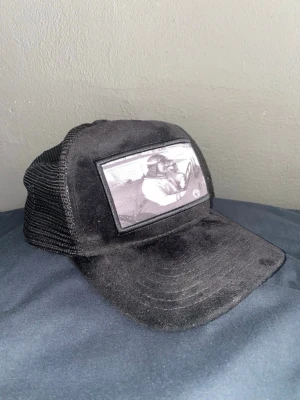 Svart meshkeps Emilio Pendenza (limited) - Svart truckerkeps från Emilio Pendenza med meshpaneler och böjd skärm. Framtill sitter en svartvit patch med motiv av en person i bil. Kepsen har justerbar baksida och logotypdetaljer på insidan. Materialet är en mix av tyg och mesh för luftig känsla.