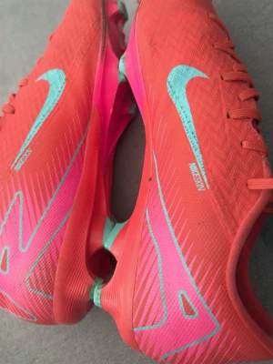 Nike Mercurial rosa fotbollsskor - Säljer ett par Nike Mercurial fotbollsskor i en rosa och röd färg med turkosa detaljer men tyvärr så är plasten på skosnören trasig logga. Skorna har en smal, aerodynamisk form och är tillverkade i syntetmaterial för bästa känsla på planen. Perfekta för dig som vill sticka ut på fotbollsplanen 