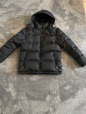 Svart pufferjacka från Polo Ralph Lauren - Snygg svart pufferjacka från Polo Ralph Lauren med huva och dragkedja framtill. Jackan har två sidofickor med dragkedja och en bröstficka med det klassiska röda Polo-logot. Perfekt för kalla vinterdagar tack vare det vadderade materialet.