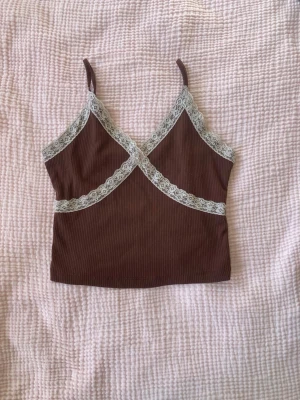 Brun ribbad linnetopp med spets - Säljer en brun croppad linnetopp med smala axelband och dekorativa vita spetsdetaljer som korsar framtill. Toppen är ribbad och har en mjuk, stretchig känsla. Perfekt för varma dagar eller att styla lager på lager.