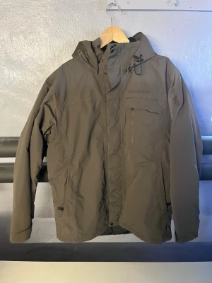 Brun vindjacka från Everest med huva - Militärgrön skidjacka från Everest med justerbar huva och dragkedja framtill. Jackan har flera fickor, inklusive en bröstficka med dragkedja, och är tillverkad i ett slitstarkt material som står emot vind och snö. Perfekt för blåsiga dagar och outdoor-äventyr.