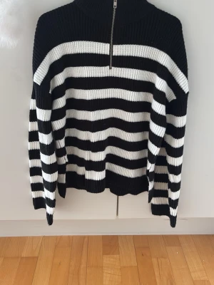Randig stickad tröja Vero Moda - Säljer en svartvit randig stickad tröja från Vero Moda. Tröjan har halv dragkedja framtill, hög krage och är ribbstickad. Perfekt för dig som gillar klassiska mönster och vill ha en mysig, stilren look.
