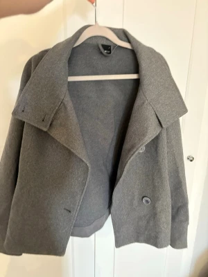 Grå kort kappa med bred krage - Snygg grå kappa med bred krage och dubbelknäppning framtill. Modellen är kortare och har en avslappnad passform. Perfekt för sommaren😍