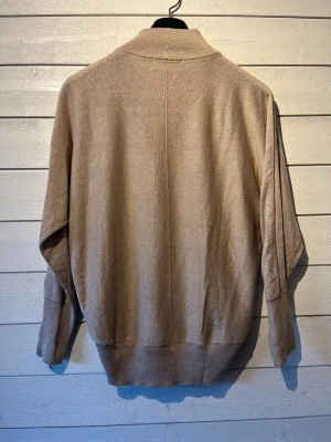 Beige m glänsande glitter trådar - In Wear  - En stilren beige stickad tröja med hög krage och långa ärmar. Tröjan har en avslappnad passform och breda ribbade muddar vid ärmslut och nederkant. Perfekt för lager på lager och enkel att matcha med olika outfits.