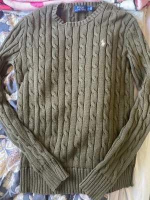 Polo Ralph Lauren - Säljer en olivgrön kabelstickad tröja från Polo Ralph Lauren. Tröjan har rund halsringning, långa ärmar och ribbade muddar. Klassisk broderad logga i vitt på bröstet. Perfekt för dig som gillar tidlös och snygg stil.