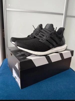 Svarta Adidas Ultraboost sneakers - Snygga svarta Adidas Ultraboost sneakers med stickad ovandel i mesh och klassiska tre ränder på sidan. Skon har en vit, responsiv Boost-sula för extra komfort och svart yttersula. Perfekt för dig som gillar stilrena och bekväma sneakers.