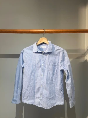 Ljusblå skjorta - Klassisk ljusblå skjorta i bomull med lång ärm och bröstficka. Skjortan har knappar framtill och en enkel, stilren design som passar till många olika looks. Perfekt för dig som gillar en clean och avslappnad stil.