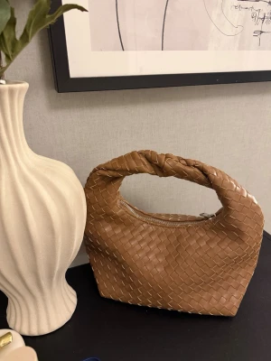 Brun flätad handväska med twistad handtag - Säljer en snygg brun handväska i flätad design med twistat handtag och dragkedja från Ginatricot. Väskan har en modern, mjuk form och är tillverkad i konstläder. Perfekt accessoar för dig som gillar trendiga detaljer och stilren look. Aldrig använd, helt nyskick. Pris går att diskutera😊
