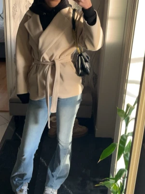 Beige omlottkappa med knytskärp - Stilren beige omlottkappa med knytskärp i midjan och bred krage från missäy. Kappan har en avslappnad passform och är tillverkad i ett mjukt tyg som ger en snygg siluett. Perfekt att slänga över en hoodie för en casual look.