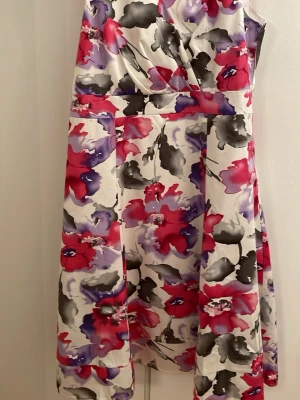 Blommig midiklänning i satin - Snygg klänning med blommigt mönster i rött, lila, svart och vitt. Klänningen har omlott upptill, markerad midja och lätt utställd kjol. Materialet är glansigt och känns som satin. Perfekt för dig som vill sticka ut med färg och mönster.