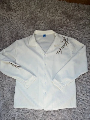 Vit skjorta med broderade detaljer - Vit skjorta i strukturerad bomull med broderade kvistar i brunt och grönt på axeln och ärmen. Klassisk krage, knäppning framtill och långärmad modell. Perfekt för dig som gillar unika detaljer och stilren design. Väldigt bra skick, rekommenderar starkt för priset. Bra till kommande säsonger också!