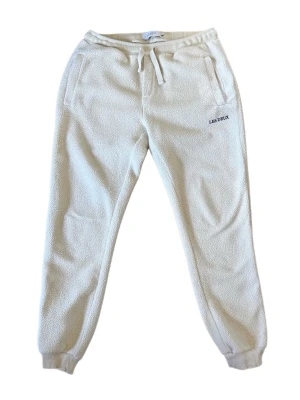 Beiga fleecebyxor från Les Deux - Mysiga beiga mjukisbyxor från Les Deux i fleece med resår och dragsko i midjan. Byxorna har ribbade muddar vid bensluten, två sidofickor och broderad logga på ena benet. Perfekta för chill dagar eller när du vill vara extra bekväm.