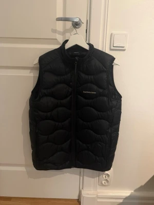 Svart dunväst från Peak Performance - Snygg svart dunväst från Peak Performance med hög krage och dragkedja framtill. Västen har ett quiltat mönster och diskret logga på bröstet. Perfekt för lager-på-lager och håller dig varm under kyliga dagar.
