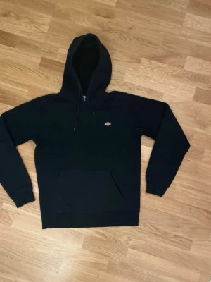 Svart hoodie från Dickies - Säljer en svart hoodie från Dickies med klassisk känguruficka och justerbar huva. Diskret Dickies-logga på bröstet. Hoodien har ribbade muddar vid ärmslut och nederkant. Perfekt för en avslappnad streetwear-look.