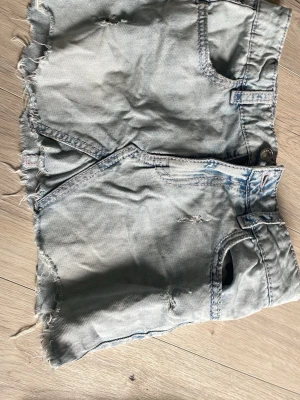 Ljusblå jeansshorts med råa kanter - Snygga ljusblå jeansshorts med råa, fransiga kanter och slitna detaljer. Shortsen har klassiska fickor fram och bak, med dekorativa sömmar i rosa på bakfickorna. Perfekta för en avslappnad sommarstil.