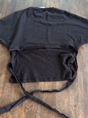 Svart croppad topp med knytband - Svart croppad topp i bomull med rund hals och korta ärmar. Toppen har långa knytband i midjan som kan knytas i ryggen. Säljer då den aldrig kom till användning 