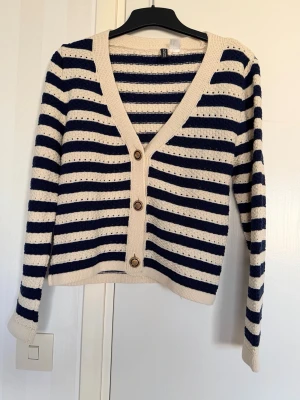 Randig kofta med knappar från H&M - Snygg kofta från H&M med marinblå och vita ränder. Koftan har v-ringning, tre bruna knappar framtill och ribbade muddar. Perfekt till jeans eller kjol för en avslappnad look.