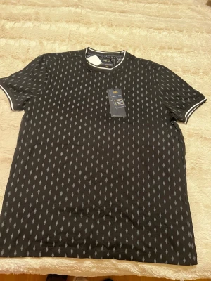 Svart mönstrad t-shirt Nickel & Dime - Svart t-shirt från Nickel & Dime med diskret grått mönster över hela tröjan. T-shirten har rund halsringning med vit kant och korta ärmar med vit detalj vid ärmslut. Tillverkad i mjuk bomull för en skön känsla.