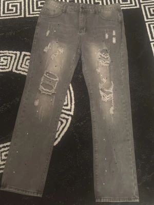 Grå slitna jeans med hål - Säljer ett par grå jeans med raka ben och tydliga slitningar samt hål på framsidan. Jeansen har en tvättad look med färgstänk och klassiska fem fickor. Perfekta för en avslappnad och trendig stil.