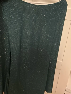 Mörkgrön glittrig långklänning - Mörkgrön långklänning med glittriga detaljer och öppen rygg med knytband. Klänningen har lång ärm och ett elegant fall baktill. Använd 1 gång sedan varit i garderoben 