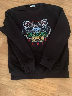 Svart Kenzo hoodie med tigerbroderi - Cool svart hoodie från Kenzo med stort färgglatt tigerbroderi och texten 'Kenzo Paris' på bröstet. Klassisk rund halsringning och ribbade muddar. Perfekt för dig som vill sticka ut med en ikonisk design. Säljer den för den blev liten efter 1 månsd