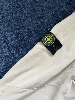 Beige hoodie från Stone Island Junior - Snygg beige hoodie från Stone Island Junior med den klassiska svarta logotypen med gul brodyr på ärmen. Hoodien är tillverkad i mjukt bomullsmaterial och har en avslappnad passform. Perfekt för en stilren och trendig look. Sitter som en Storlek S Herr